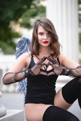 Portrait of a beautiful girl with red lips in a black bodysuit, gloves and blue wings. Posing sitting in front of the columns. Shows heart with hands