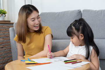 Asian mother look at her daughter painting on white paper on the wooden table with beautiful smile and pride.