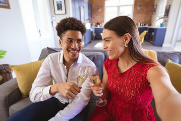 Happy young biracial couple toasting champagne flutes while taking selfie at home - Powered by Adobe