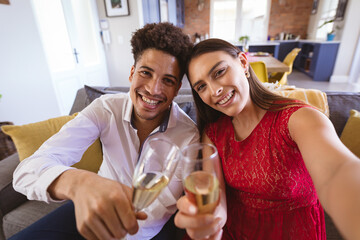 Portrait of smiling young biracial couple with champagne flutes taking selfie at home during party - Powered by Adobe