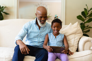 Smiling african american senior man with granddaughter using digital tablet while sitting on sofa