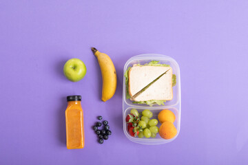 Fresh fruits and juice bottle by sandwich in tiffin box on purple background with copy space