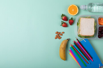 Fruits with sandwich in tiffin box by water bottle, color pencils and books on green background