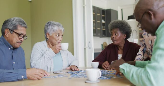 Group of happy diverse senior friends drinking coffee and doing puzzle at home