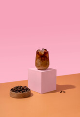 Ice coffee in a tall glass with cream poured over and coffee beans on pink background.