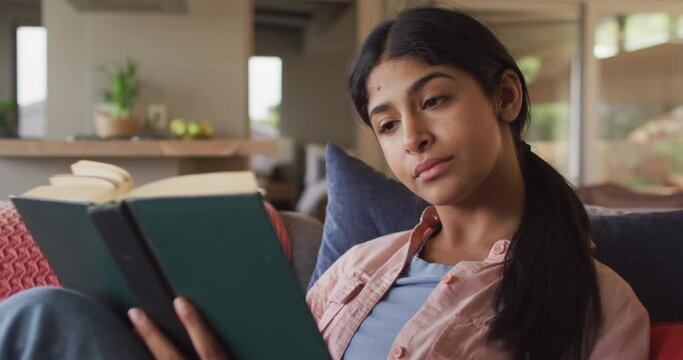 Video of biracial woman sitting on sofa and reading book