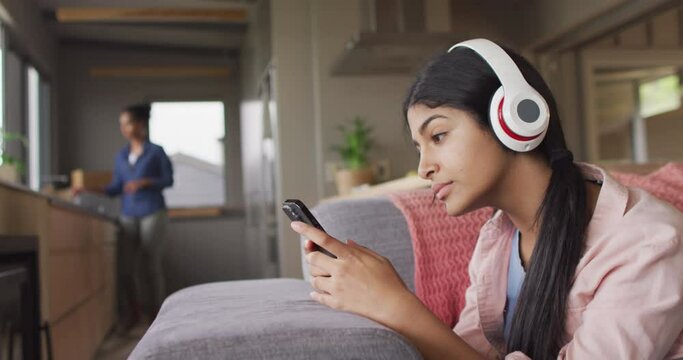 Video of happy biracial woman lying on sofa with headphones and smartphone