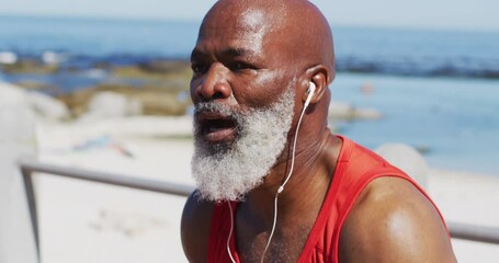 Tired senior african american man wearing headphones taking a break from running on promenade - Powered by Adobe