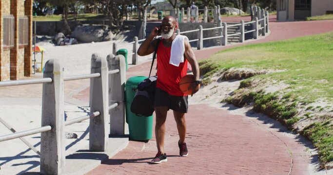 Senior african american man with basketball talking on smartphone while walking near the beach