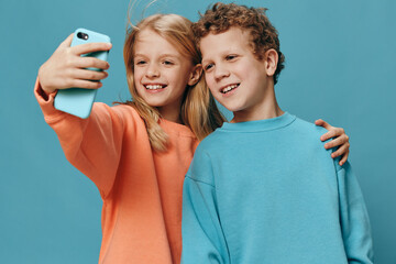 cute, beautiful children, brother and sister stand in bright clothes on a blue background and the girl takes a selfie on her smartphone hugging her brother
