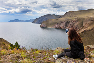 A tourist girl admires a beautiful view of Baikal Lake from a height of a coastal rocks and takes...