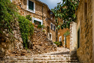 Narrow uphill alley named Carrer Església with curved cobbled steps in the idyllic mediterranean mountain village Fornalutx lined with mediterranean stone houses and overgrown walls at Mallorca.