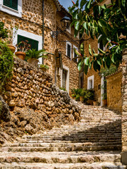 A narrow curved alley with cobbled steps called Carrer de l'Església leads through the rural Mallorca mountain village Fornalutx with mediterranean stone houses and overgrown walls at a summer day.
