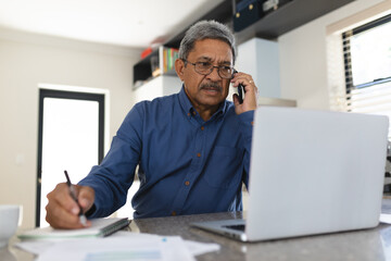 Mixed race man sitting in kitchen talking on smartphone and using laptop