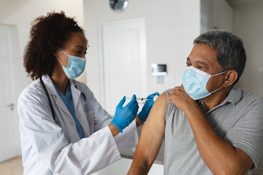 Mixed race female doctor wearing mask giving vaccination to senior man at home