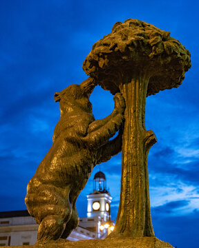 Statue of the Bear and the Strawberry Tree in the Puerta del Sol square in the city of Madrid with the clock chimes in the background during a sunset on a cloudy autumn day