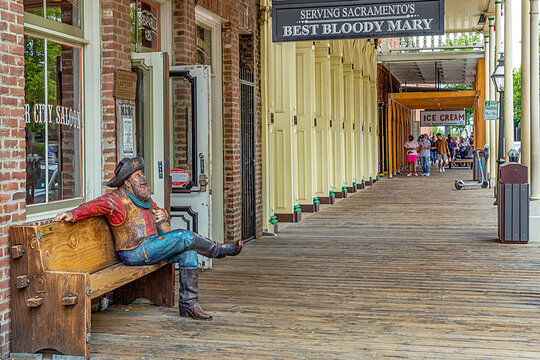 wooden statue of cowboy sitten on a bench in the old town of Sacramento with smoking pipe in front of a bar and tlurists in background at historic sidewalk