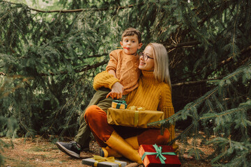 Stylish mother and son in yellow sweaters with Christmas gifts sit near spruce tree