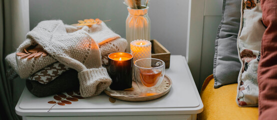 Autumn still life concept. Cozy autumn composition with burning candles, glass cup of tea, stack warm sweaters on bedtable in bedroom.
