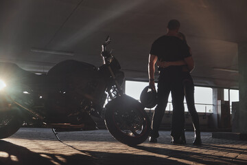 Young couple hugging standing by on a motorcycle in underground parking garage On the Sunset
