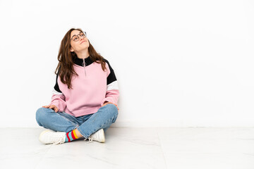 Fototapeta premium Young caucasian woman sitting on the floor isolated on white background looking up while smiling