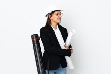 Young architect caucasian woman with helmet and holding blueprints over isolated background looking to the side