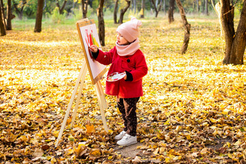 A little girl in a red coat paints a watercolor on the easel in the park against the background of the autumn landscape