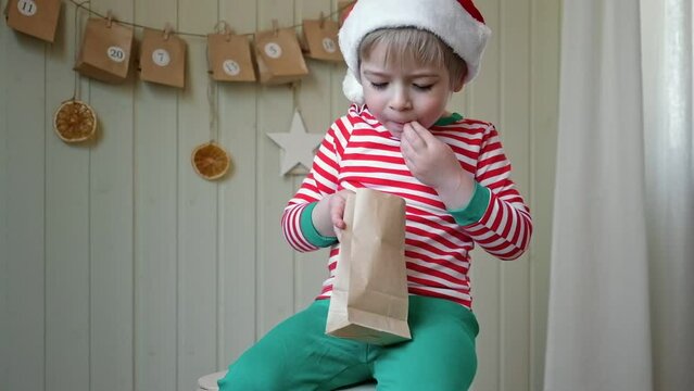 Boy in pajamas, Santa cap eating sweet candy on paper advent calendar with presents background. Little happy child with gift of handmade Christmas calendar on wall. Celebrating at home Xmas tradition.