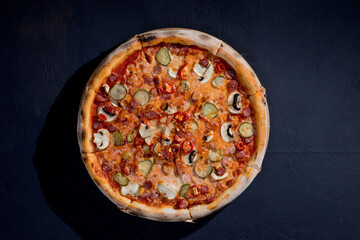 Appetizing pizza with hunting sausages, mozzarella cheese, pickled cucumbers, chili peppers on a tomato base on black background