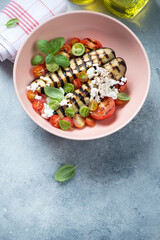 Roseate bowl with grilled eggplant, goat cheese and mini tomatoes, vertical shot on a light-blue stone background with space, high angle view