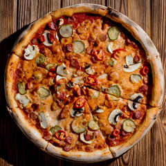 Appetizing pizza with hunting sausages, mozzarella cheese, pickled cucumbers, chili peppers on a tomato base on wood background