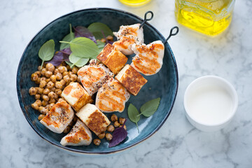 Haloumi and chicken skewers with roasted chickpeas in a blue bowl, elevated view on a light-grey marble background, horizontal shot
