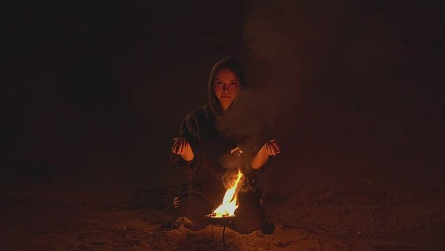 a girl in a hood holds sand in front of a bowl of fire