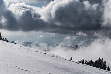 Winter landscape. Dramatic sky with grey clouds above snow covered steep mountain slope. Untouched white powder snow contrasts the grey clouds.