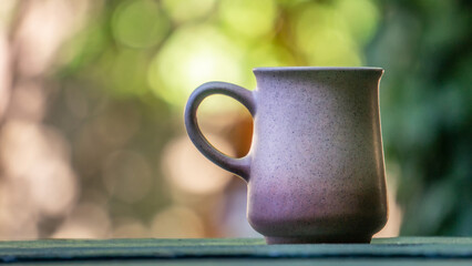 Ceramic tea cup on green wooden table. Blurry background with outdoor garden, plants and vegetation. Hot beverage mug and warm tones garden bokeh.