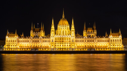 Naklejka premium Panorama of the Hungarian Parliament building at night. Night view of the illuminated main facade. Warm lights reflected in the water. Situated in Budapest on the eastern bank of the Danube.