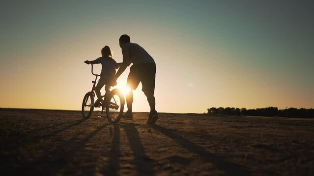 Happy family concept. Dad teaches daughter to ride bicycle on green grass. Kid rides a bicycle along rural road. Green energy.Dad teaches his daughter to ride bike for the first time in park at sunset