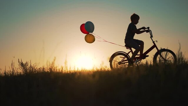 Dream child. Silhouette of kid on bike in park. Boy rides in park on green grass. Child games in nature.Traveling with balloons on bike.Active child freedom in summer.Boy learn to ride bike in nature.