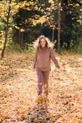 Young woman in hoodie sweater kicks up leaves in autumn park. Sunny weather. Fall season.
