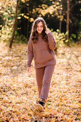 Young woman in hoodie sweater kicks up leaves in autumn park. Sunny weather. Fall season.