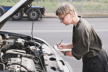 Middle-aged woman holding a notepad stands in front of a car with the hood up. Insurance agent, middle-aged woman, conducts pre-insurance inspection of car.