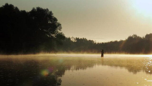 Silhouette of a fisherman on the river on a foggy summer morning. Dawn. Beautiful landscape with a river in a foggy morning