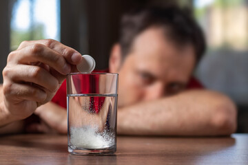 A man takes a cure for a hangover at home, throws a second fizzy pill into a glass, focusing on the hand with the pill