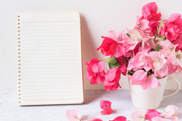 Notebook and Pink Bougainvillea flower in coffee cup on white wooden table with cement wall background