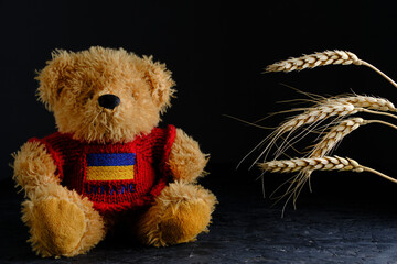 Soft toy and ears wheat on black background, bear in red jacket with flag Ukraine depicted on it.