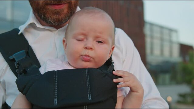 Close-up portrait of funny crying newborn baby in a slings carrier. Young father holding a child on street. Ergo backpack. Parents. Slow motion