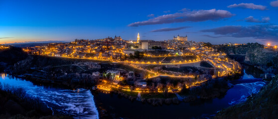 Panoramic view of the city of Toledo with the streetlights on, illuminating the streets and famous buildings, such as the Cathedral, along the Tagus River at dusk