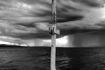 In the foreground a wooden pole, in the background an impetuous sea and a very cloudy sky, a violent downpour of rain on the horizon is clearly recognizable. Black and white photo. - obrazy, fototapety, plakaty