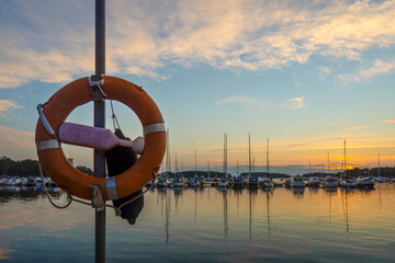 Lifebuoy against the sunset and moored boats in Syväraumanlahti - sea bay, one of the largest marinas in Rauma, Finland. Orange ring used to help people drowning in port.