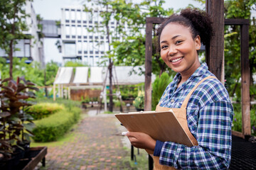 Fototapeta premium African American woman owner an ornamental garden using a tablet to check the plants on a shelf in a green house for sell. Portrait of charming caucasian woman owner of garden center